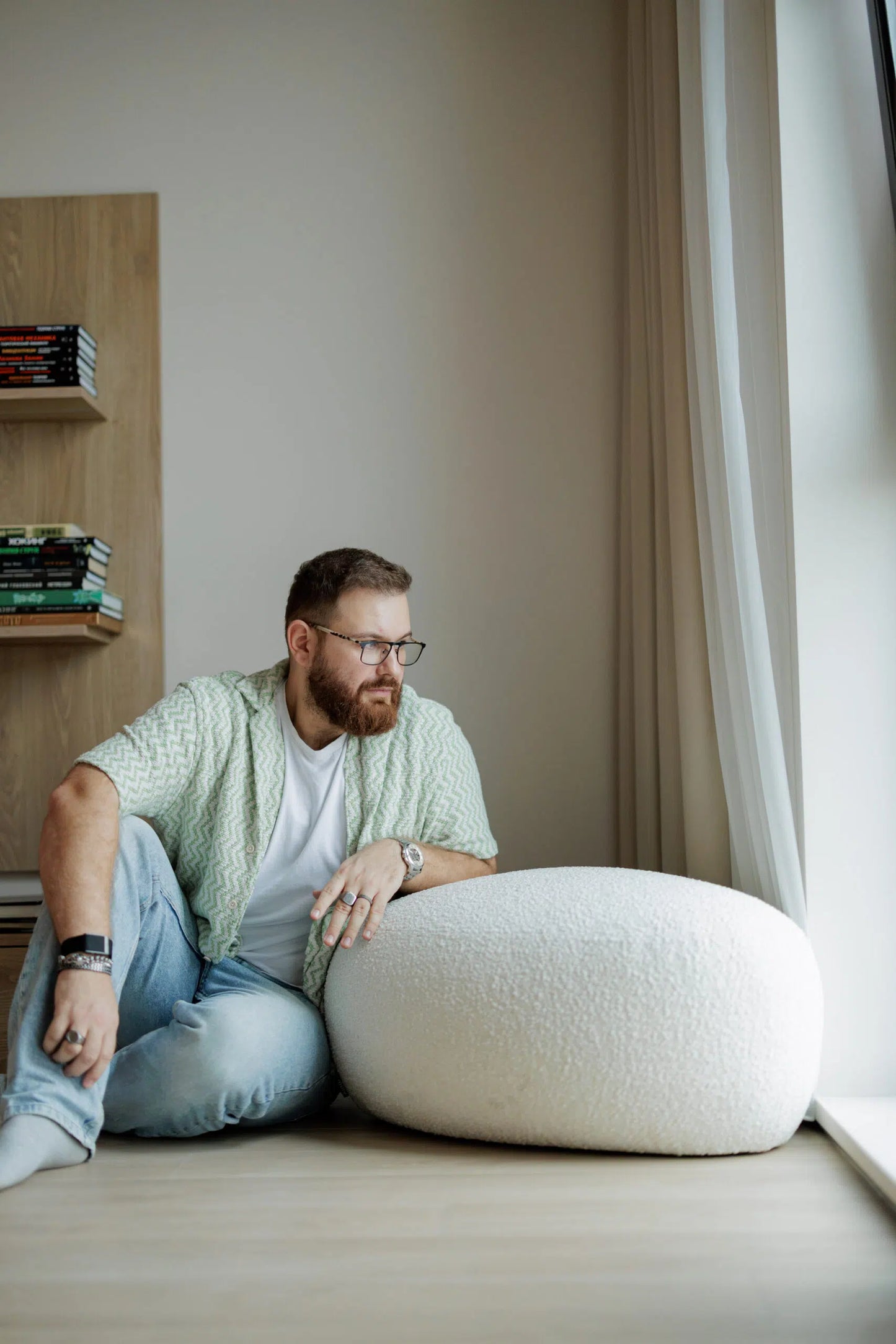 Man sitting on a white cushion in a room with a bookshelf and window.