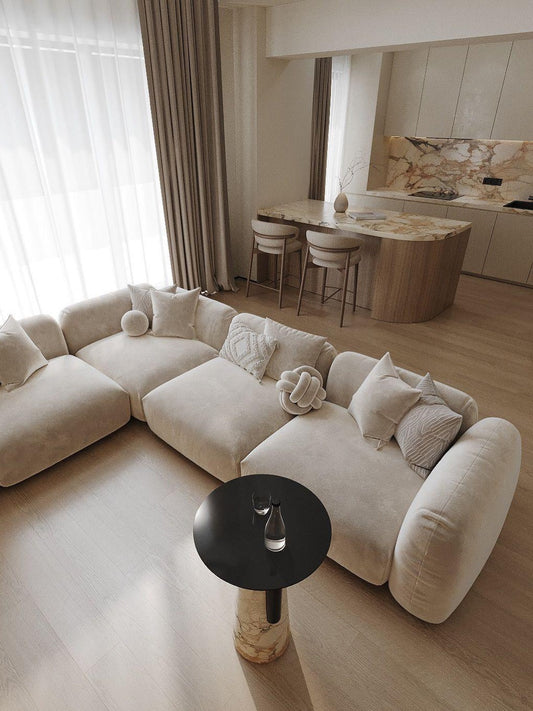Beige modulsoffa i sammet med puffeffekt och dekorativa kuddar i ett elegant, modernt beige vardagsrum med stenkök och stenbord.