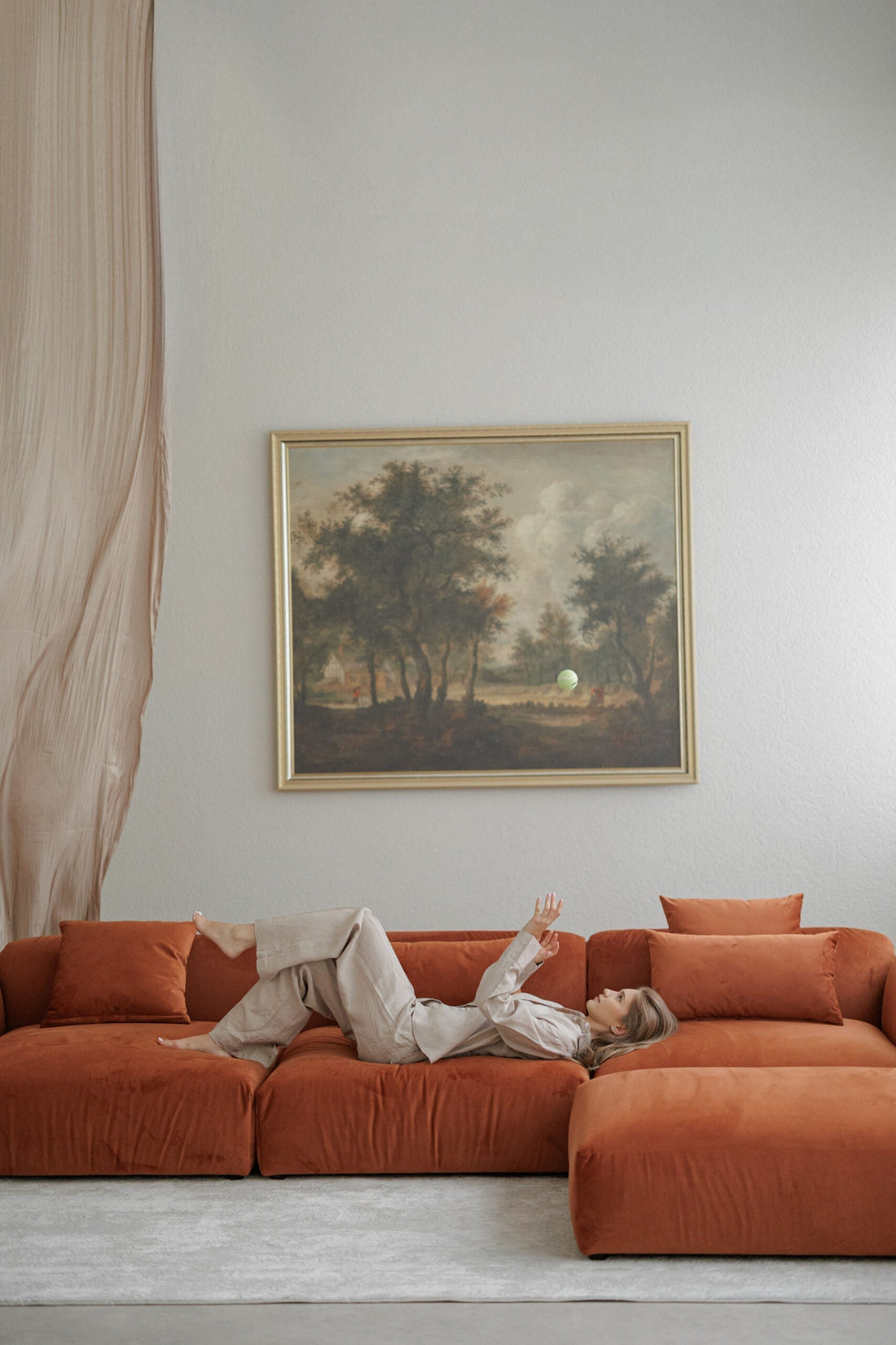 Orange velvet modular sofa with puffy effect in a studio with painting, curtains and model playing ball on it. 