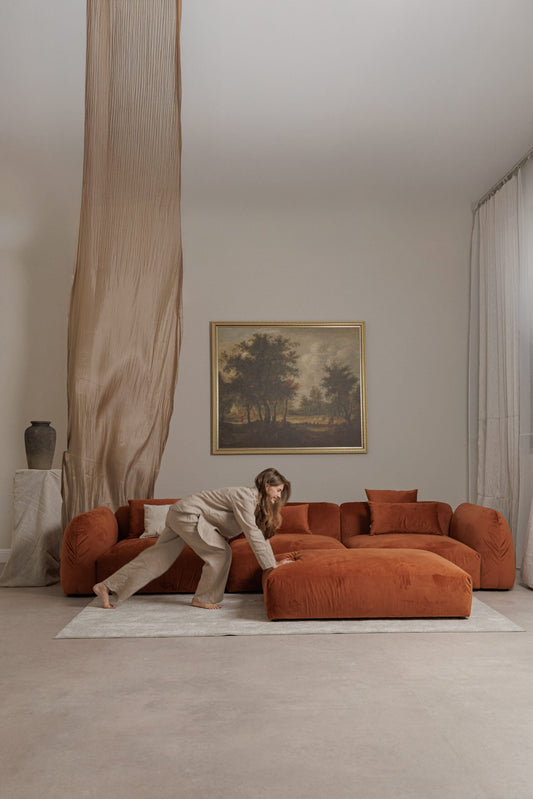 Orange velvet sofa with ottoman in a beige classic studio with painting, curtains and model moving ottoman. 