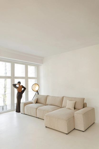 Beige sectional sofa bed  in a minimalistic living room with a person standing by a window.