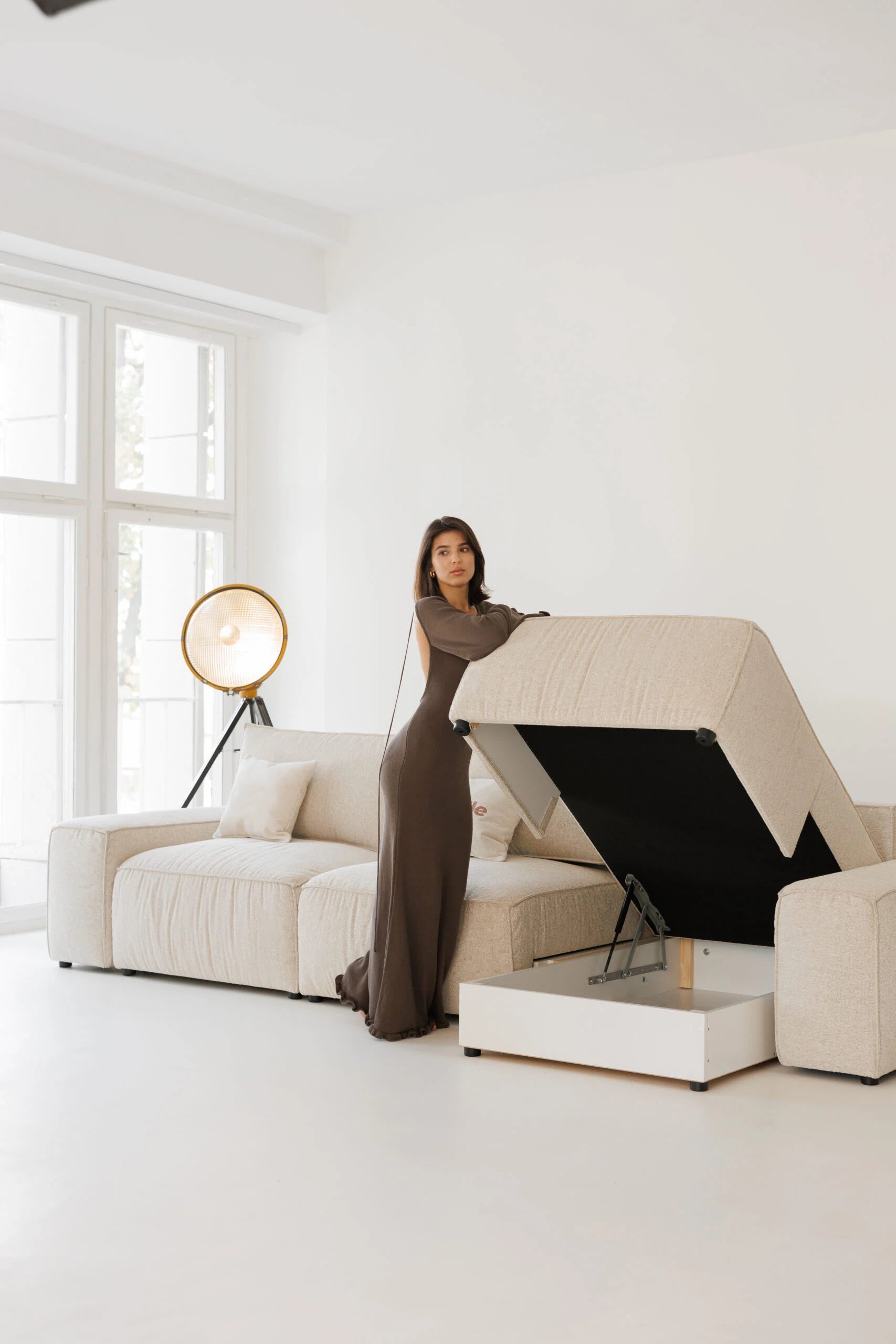 Woman in a long dress standing next to a modern beige sofa bed with storage
