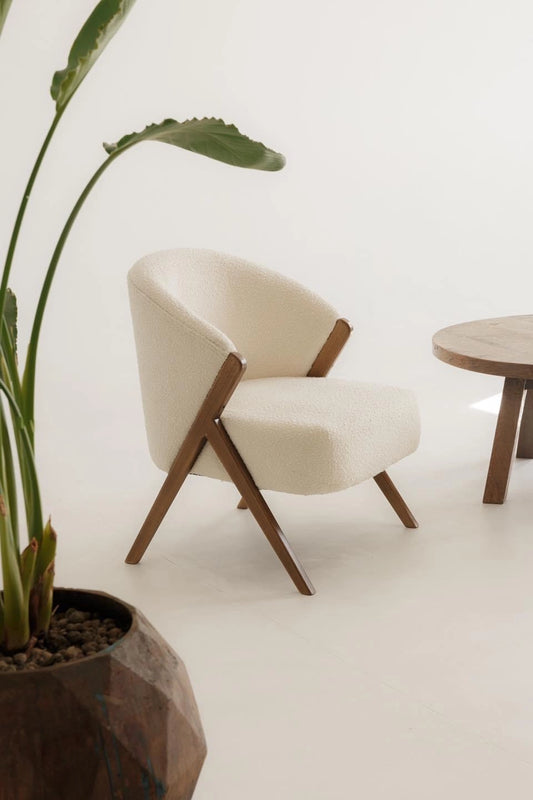 Modern white bouclé armchair with wooden arms next to a wooden coffee table and plant. 
