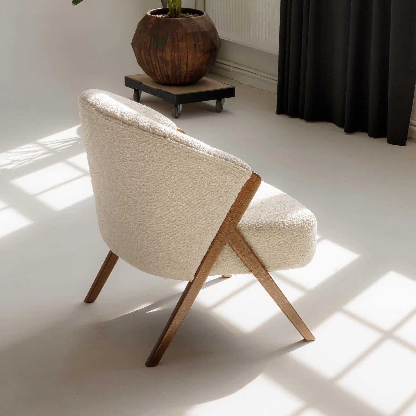 Modern white bouclé armchair with wooden arms next to a plant in a studio. 