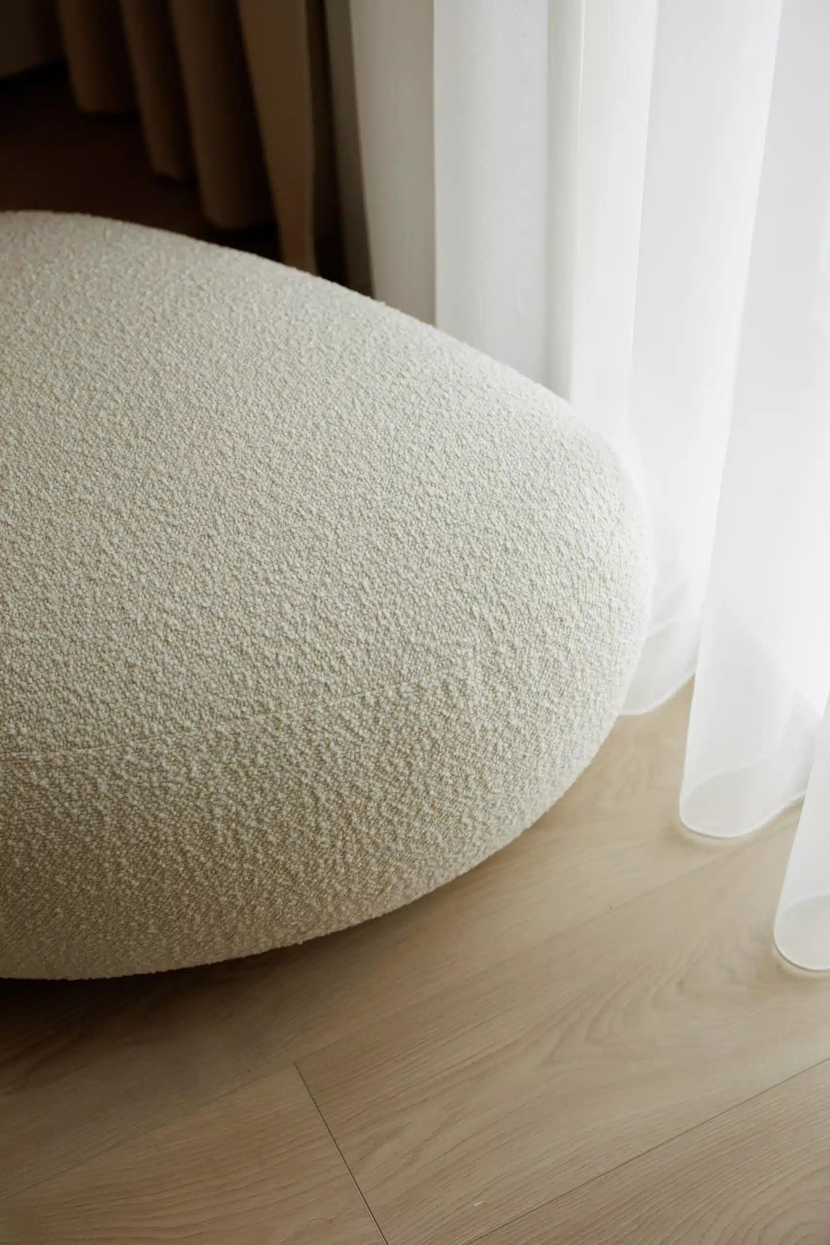 Round textured ottoman on a wooden floor with white curtains in the background