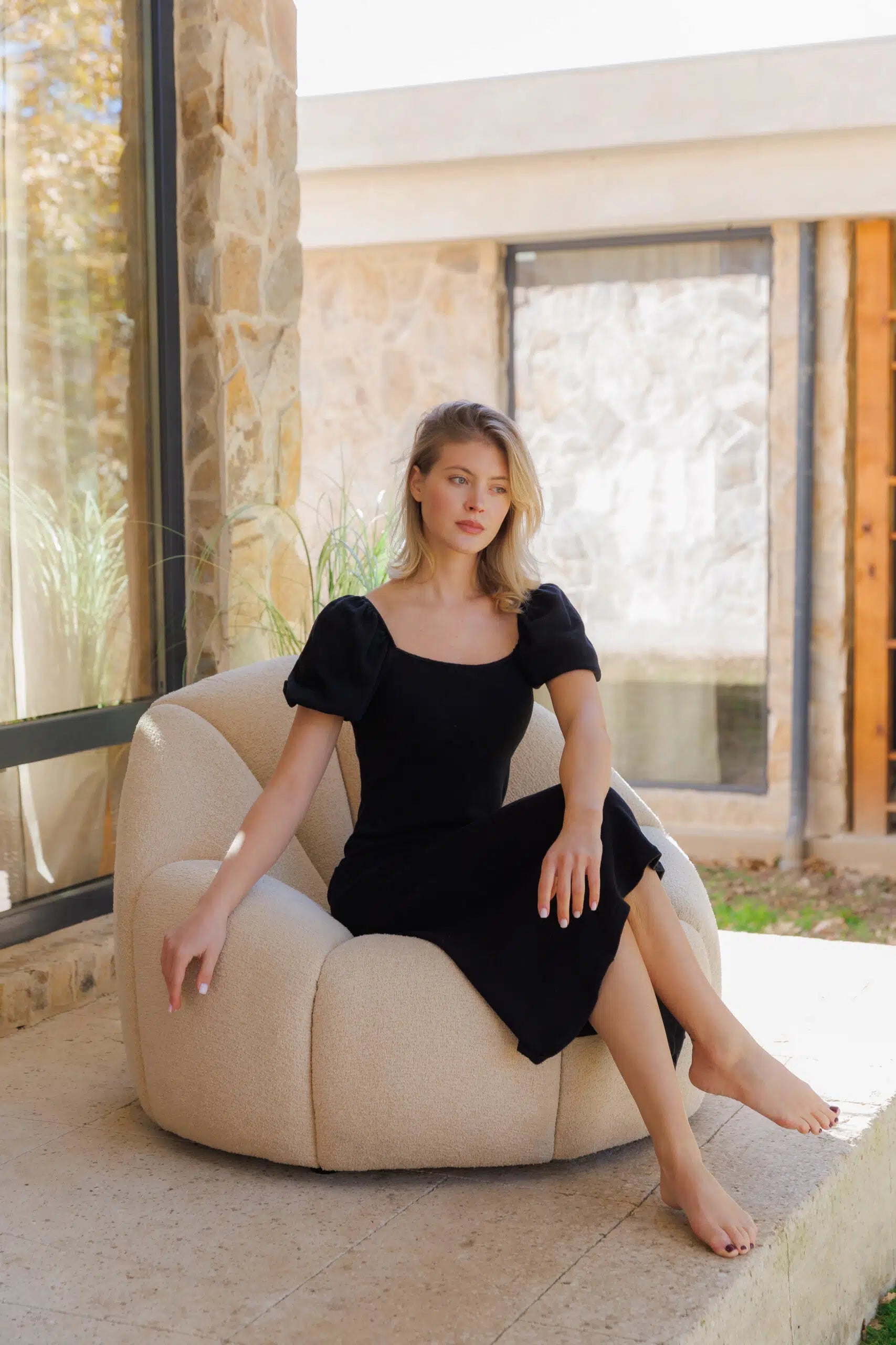 Model in a black dress sitting on a white upholstered armchair in a modern outdoor setting.