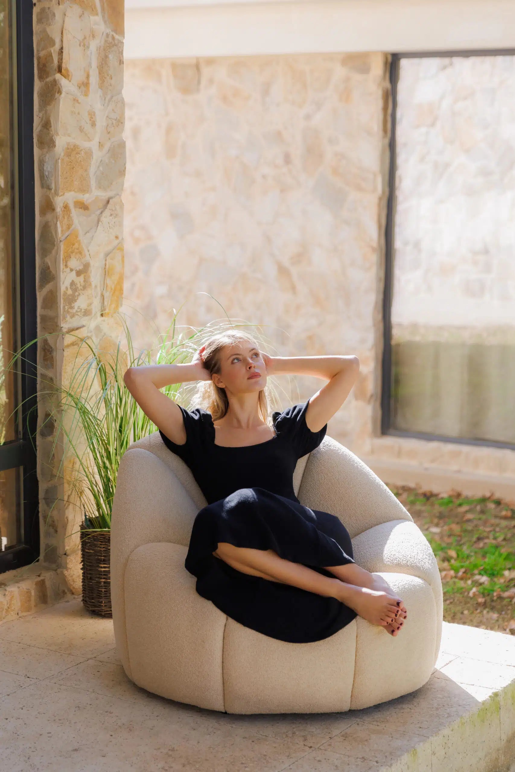 Woman sitting on a beige designer armchair in an outdoor setting with stone walls and plants.