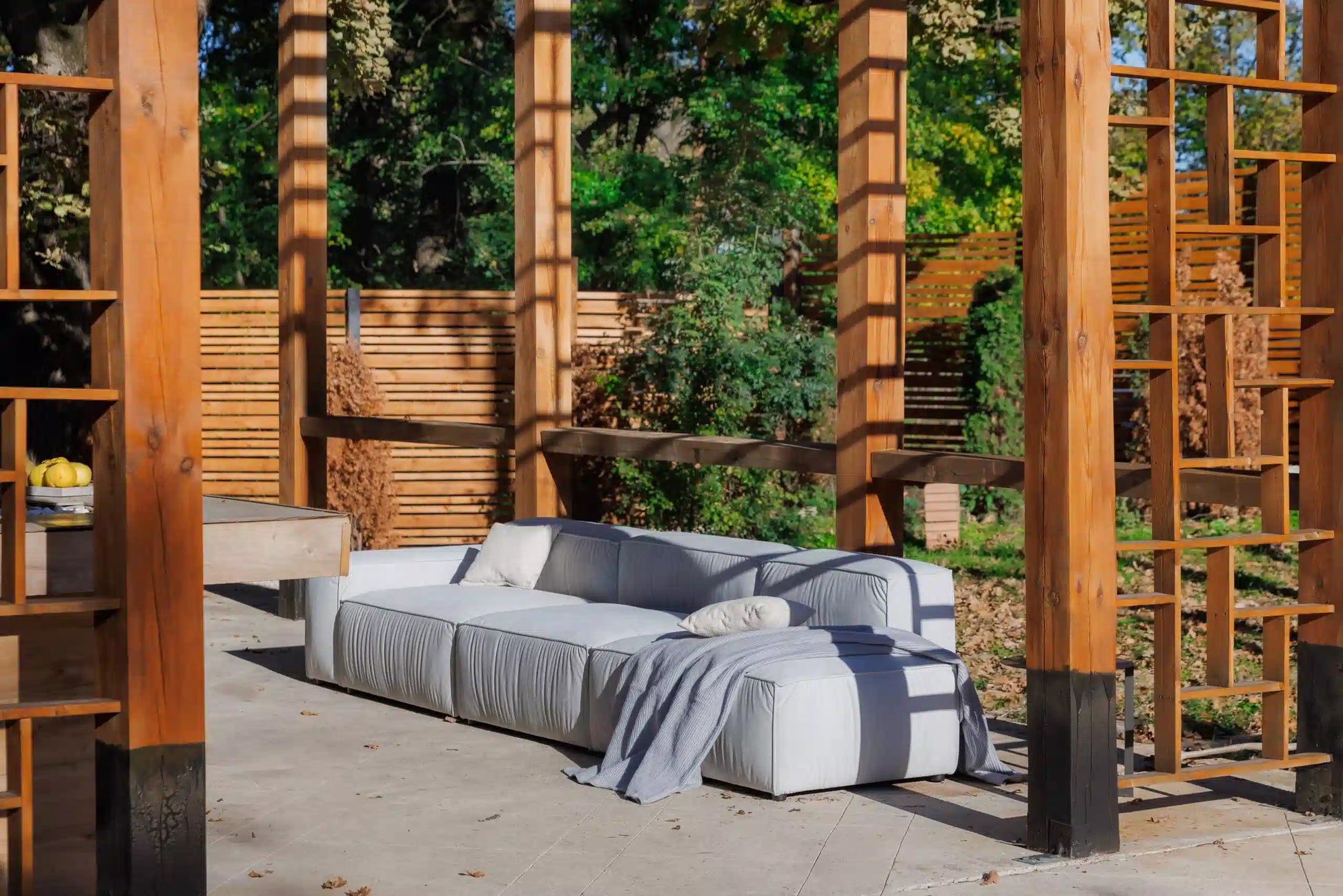 Gray luxury modular sofa with cushions on a wooden deck surrounded by wooden structures and greenery.