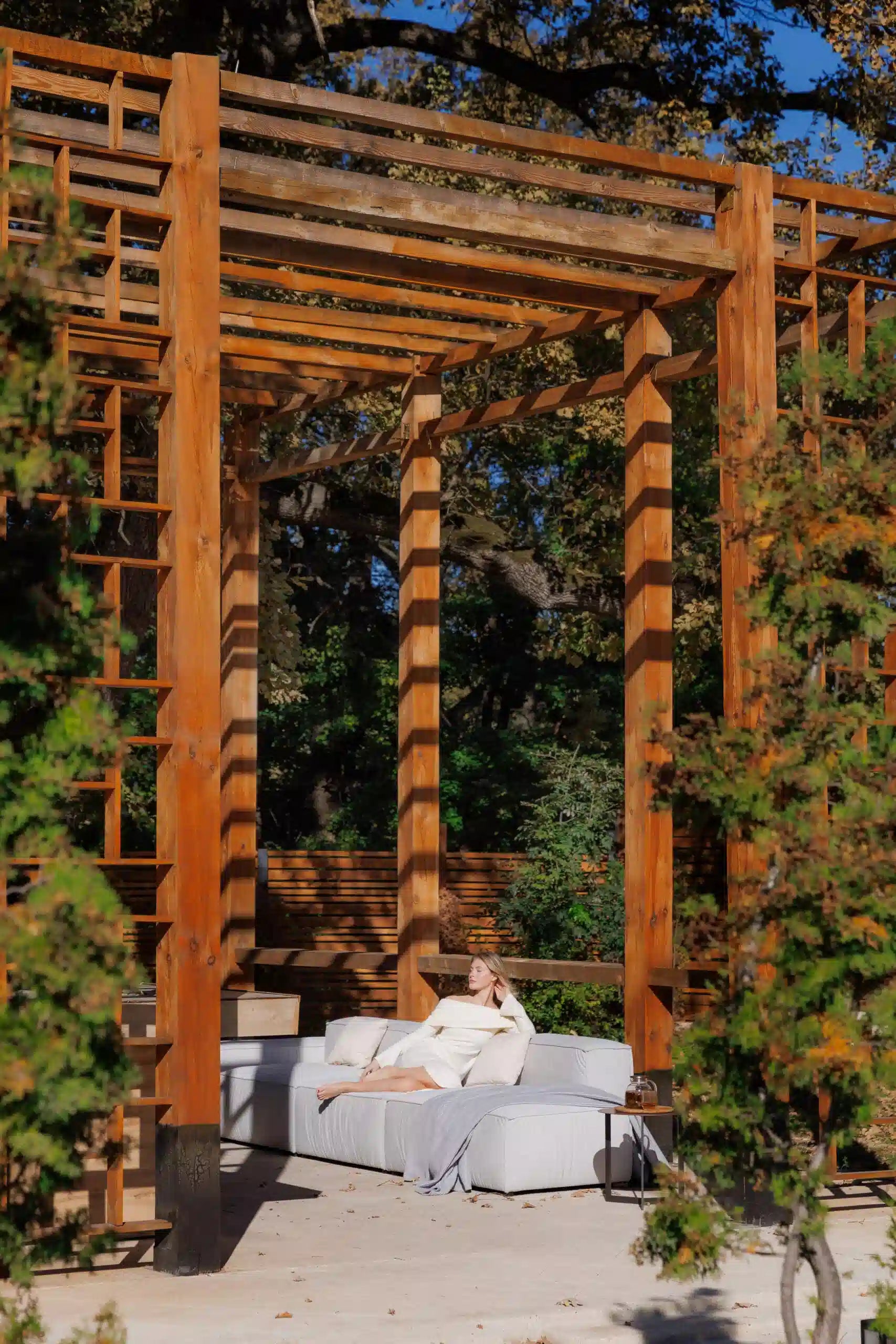 Person lounging on a luxury sofa under a wooden pergola in a garden setting
