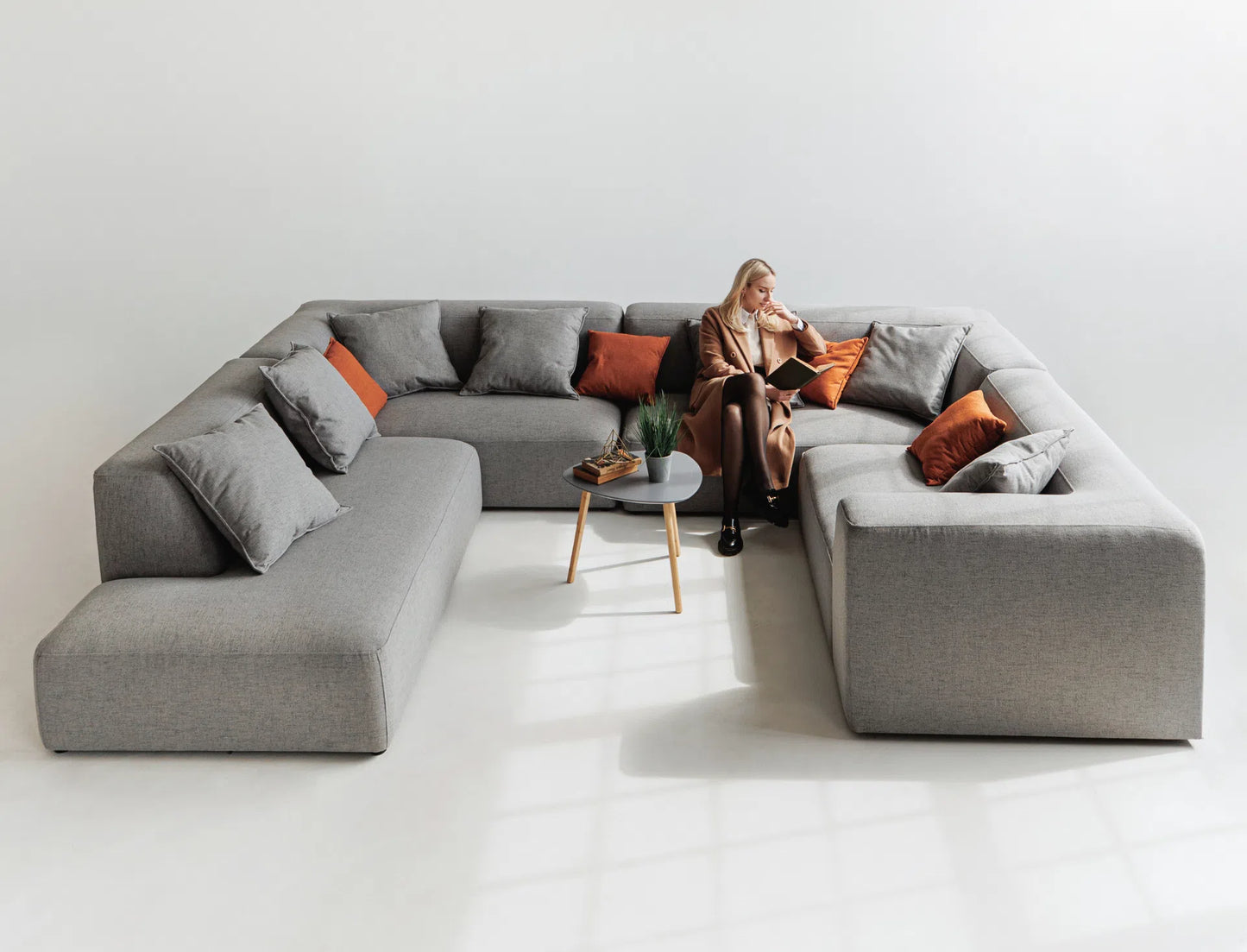 Person sitting on a large gray sectional sofa with orange cushions in a minimalistic room.