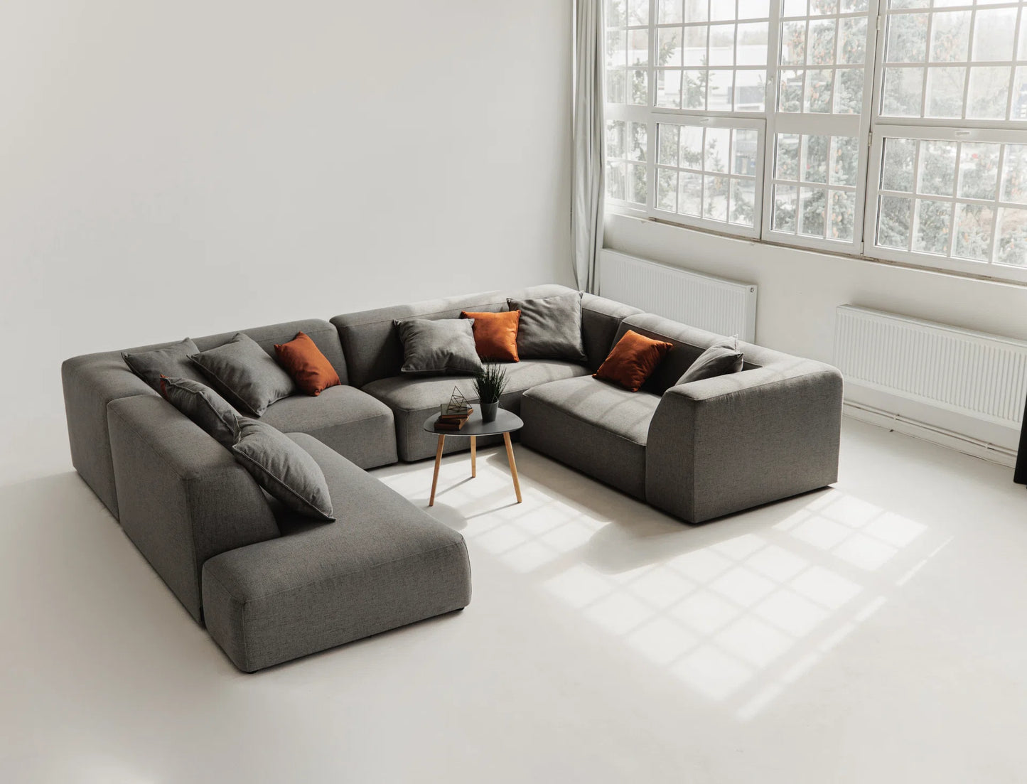 Gray sectional sofa with orange cushions in a bright room with large windows.