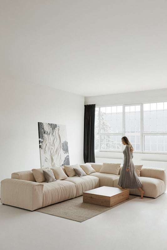 Woman standing in a modern living room with a large l shaped sofa and a painting on the wall.