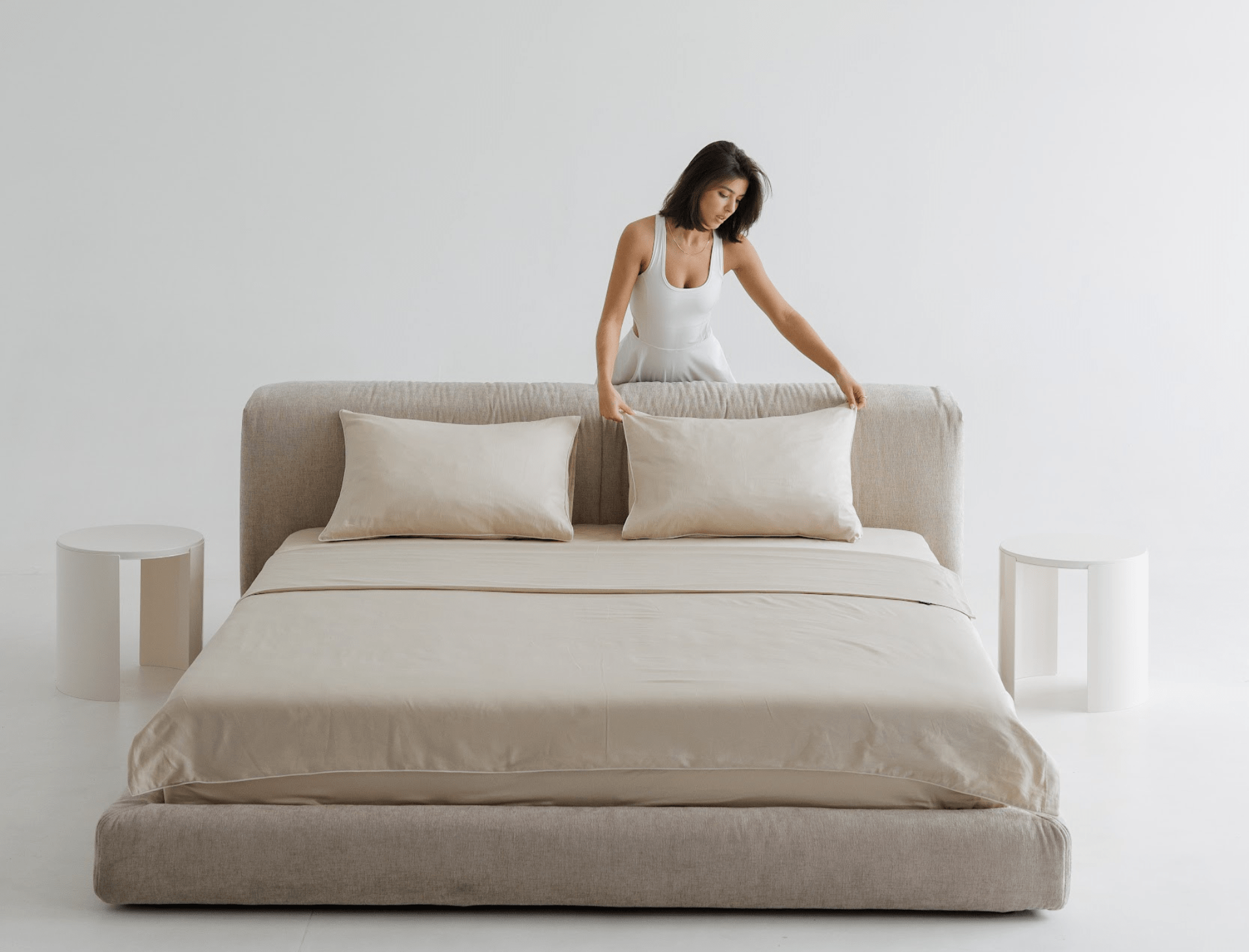 Woman arranging pillows on a beige bed in a minimalistic room.