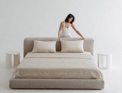 Woman arranging pillows on a beige bed in a minimalistic room.