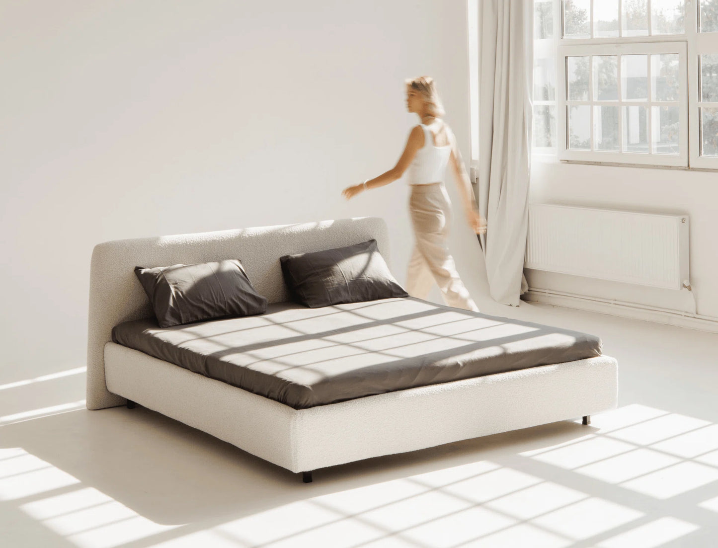 Modern white bed with dark bedding in a bright room with a woman walking by.