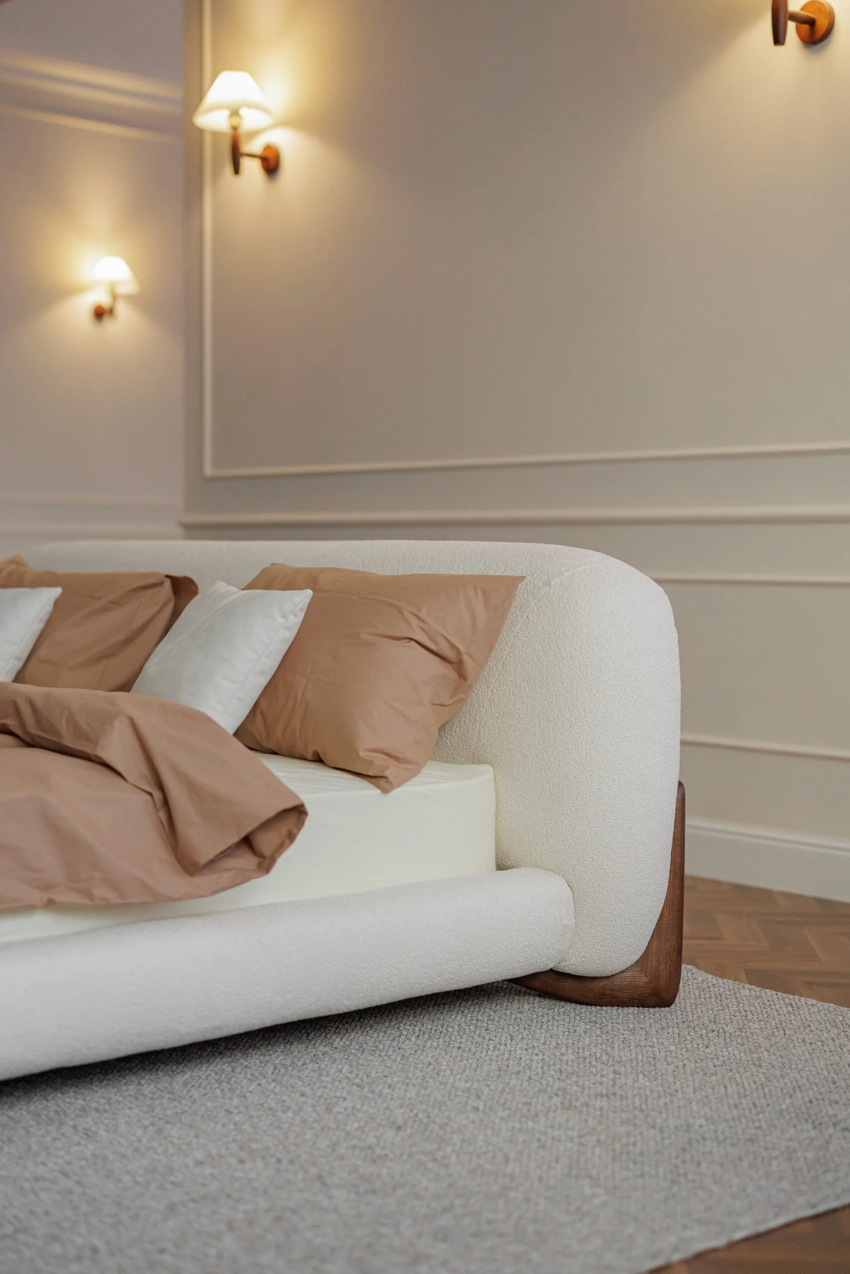 White bed with brown pillows in a room with beige walls and wall lights.