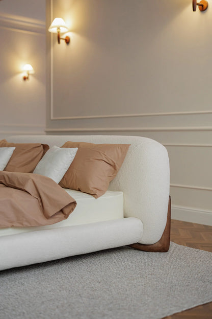 White bed with brown pillows in a room with beige walls and wall lights.