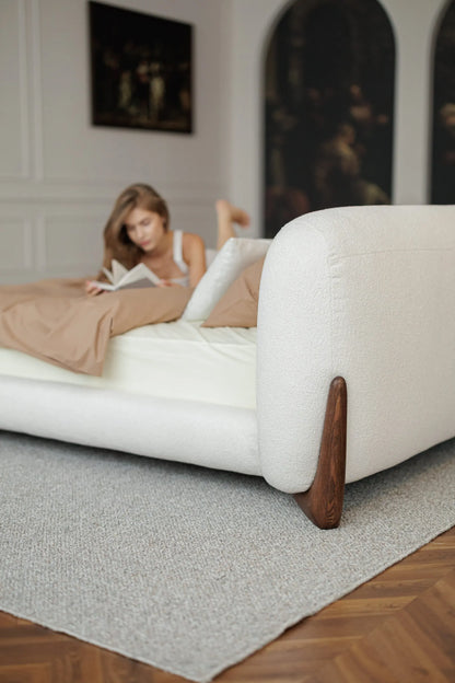 Woman lying on a large white bed with wooden legs in a room with a painting on the wall.