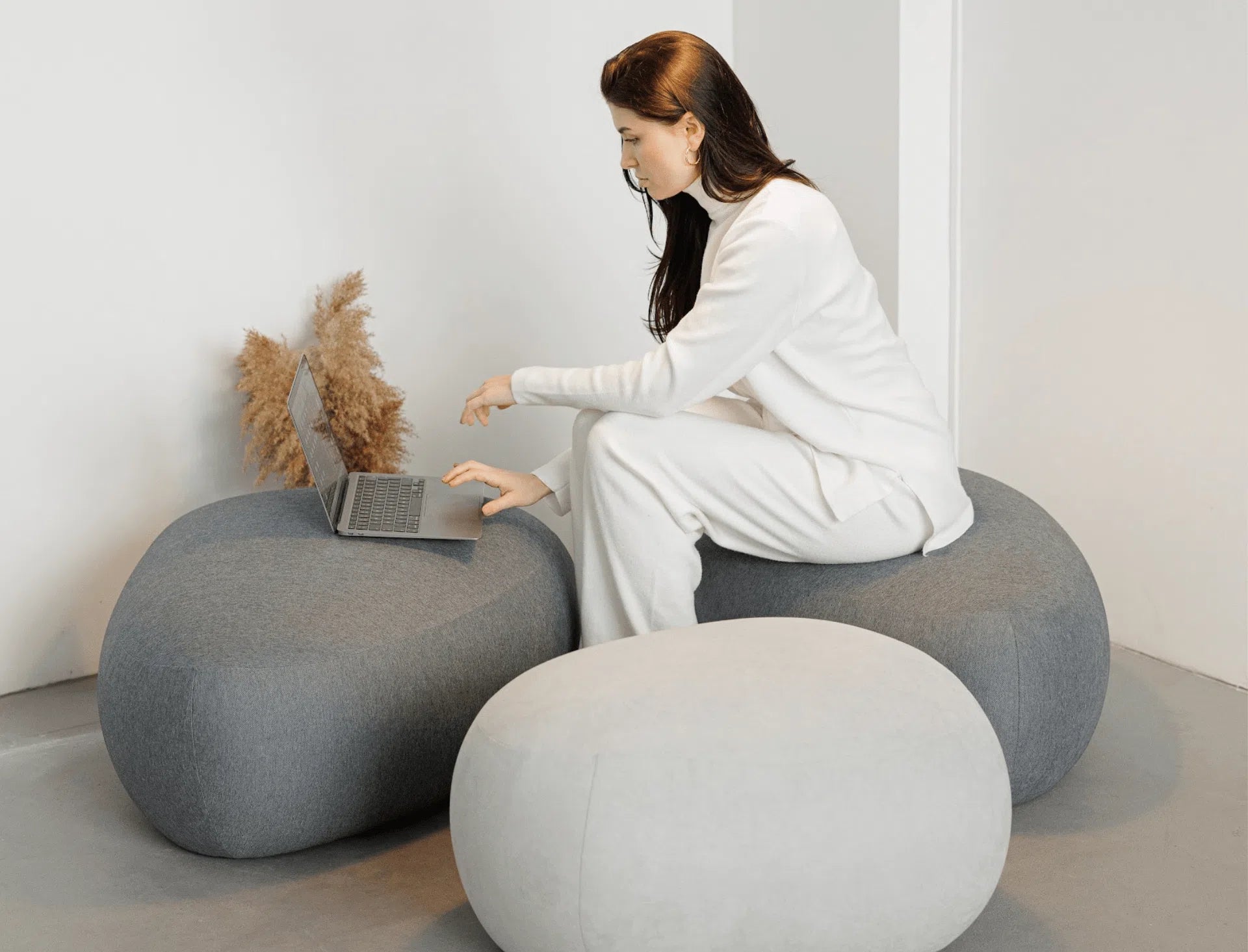 Woman using a laptop on a modern, round gray ottomans in a minimalistic room.