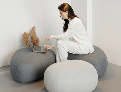 Woman using a laptop on a modern, round gray ottomans in a minimalistic room.
