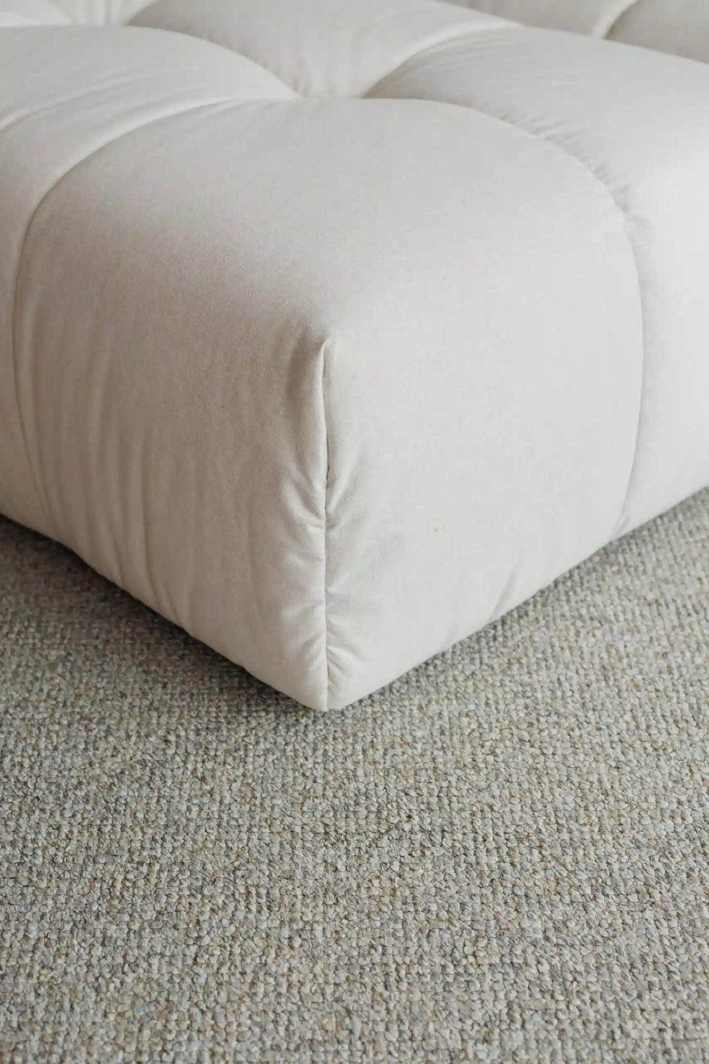 Close-up of a beige upholstered ottoman on a textured gray carpet