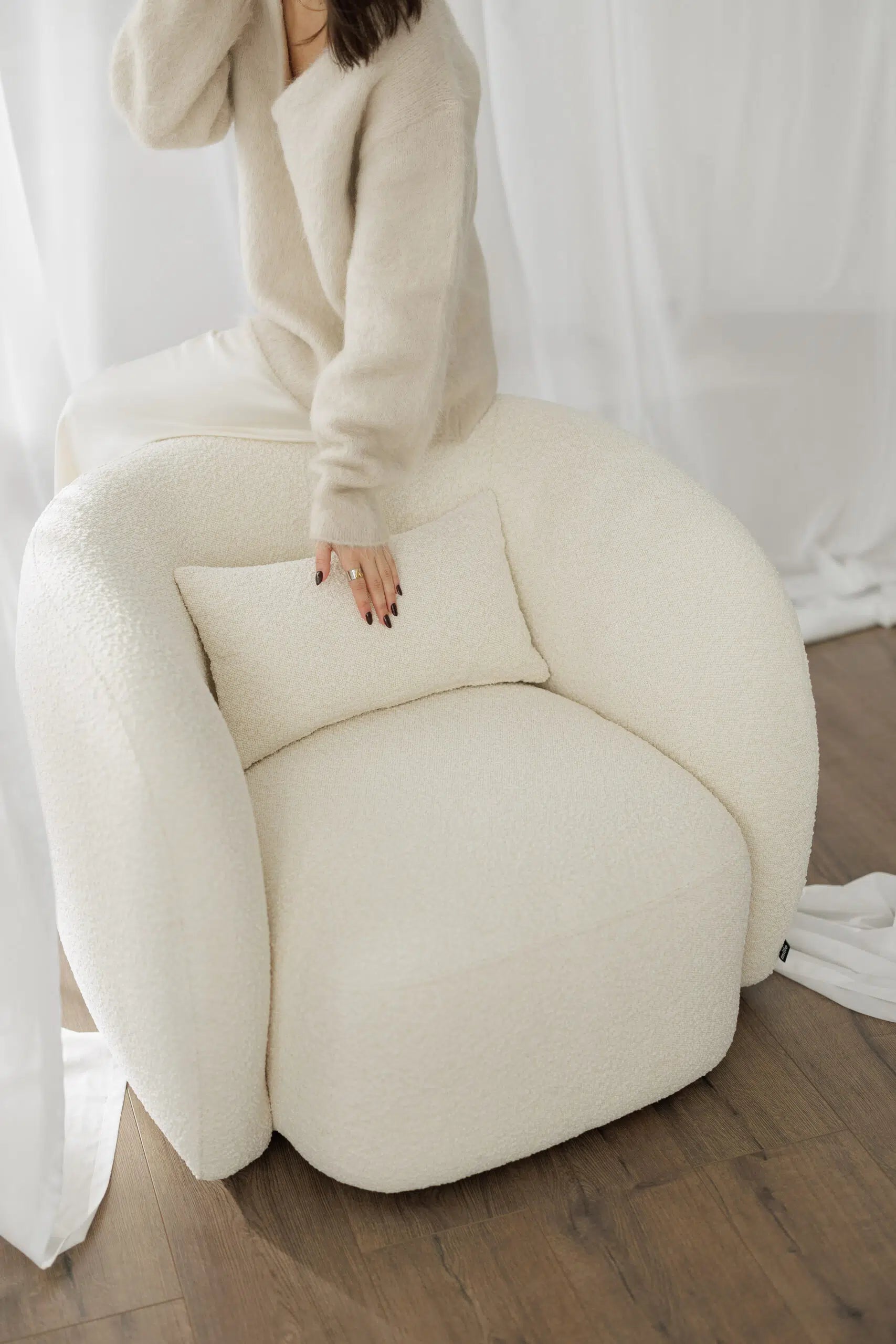 Person sitting on a large white bouclé armchair against a white curtain background