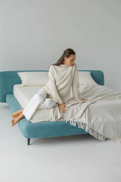 Woman sitting on a blue upholstered bed with white cushions and blanket in a minimalistic room.